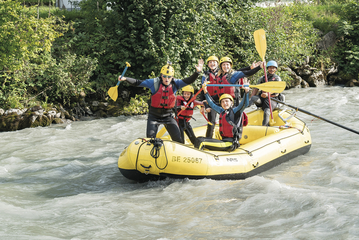 A smiling family stands on the raft with their hands in the air as they float down the river during the Family Rafting trip